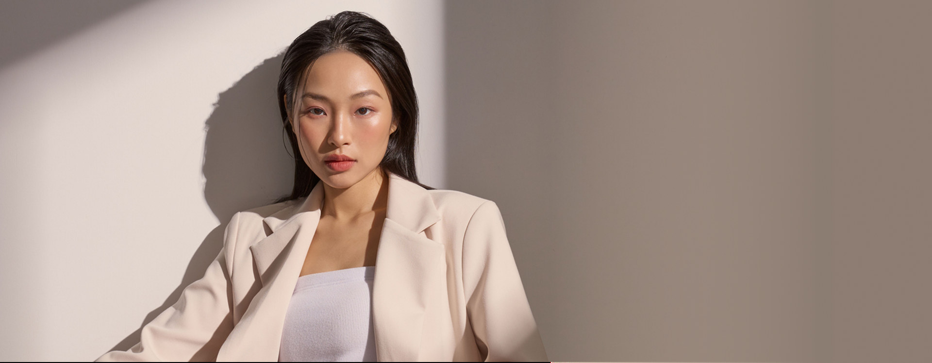 A woman in a beige blazer against a sunlit wall.