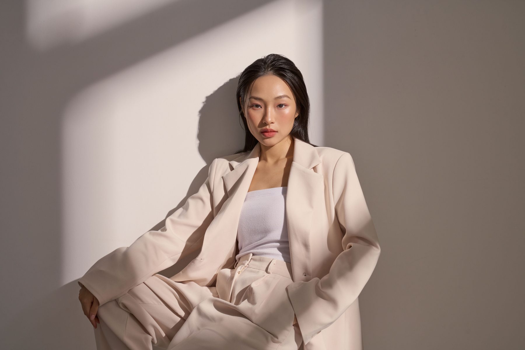 A figure in a beige suit standing against a sunlit wall.