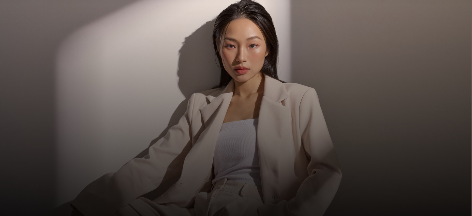 A seated figure in a beige suit against a sunlit wall.